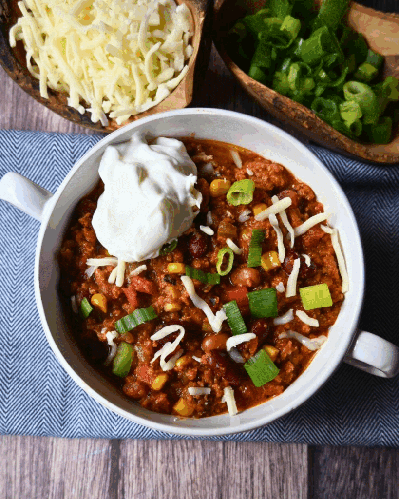 A bowl of chili with ground turkey, beans, corn, tomatoes, topped with shredded cheese and sliced green onions, sits on a wooden surface with extra cheese and onions scattered around.