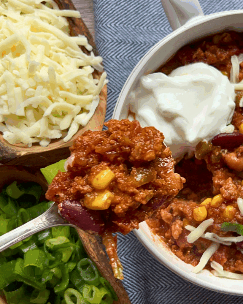 A close-up of chili in a white bowl topped with sour cream, with a spoonful of chili held above. Surrounding the bowl are bowls of shredded cheese and chopped green onions.