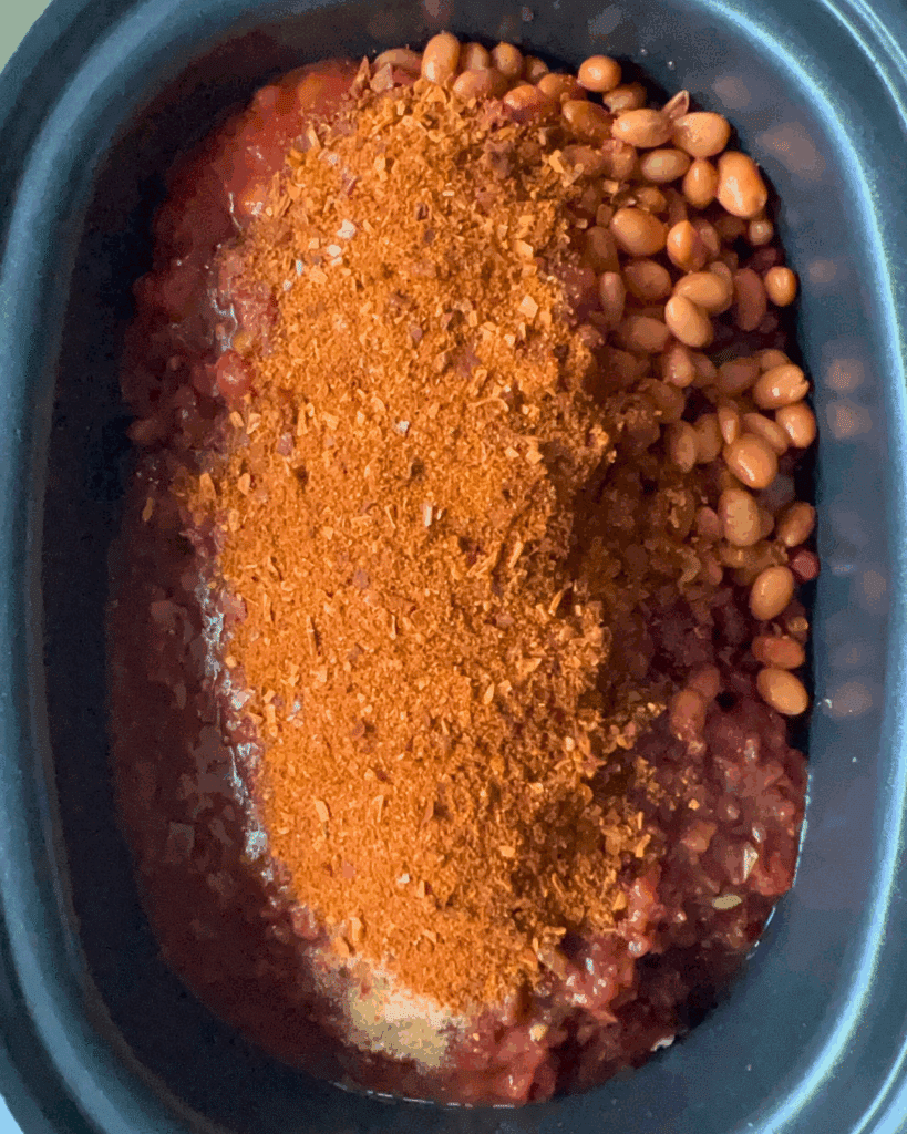 A slow cooker filled with pinto beans, diced tomatoes, and a large pile of chili seasoning, all ready to be mixed and cooked.