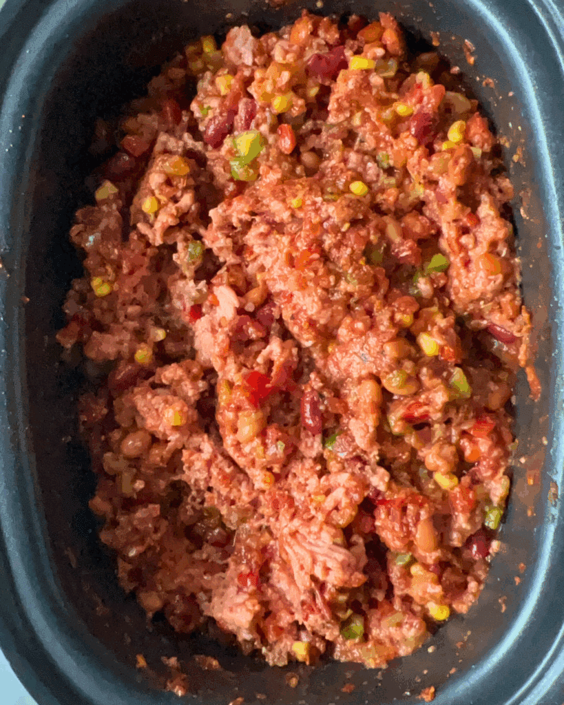 A close-up of a slow cooker filled with homemade chili, showing ground meat, red kidney beans, corn, diced tomatoes, and chopped green bell peppers mixed together.