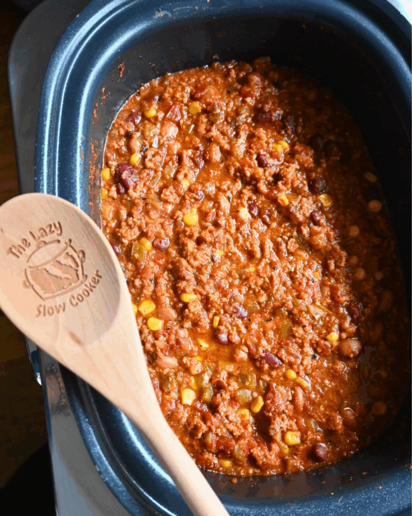 A slow cooker filled with hearty chili containing ground meat, beans, corn, and tomatoes. A wooden spoon labeled The Lazy Slow Cooker rests on the edge.