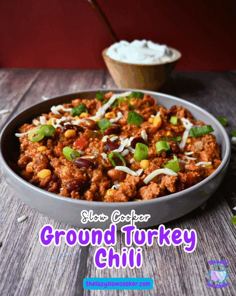 A bowl of chili topped with shredded cheese and chopped green onions sits on a wooden surface. A small bowl of sour cream is in the background. The chili contains beans, corn, ground turkey, and tomatoes.