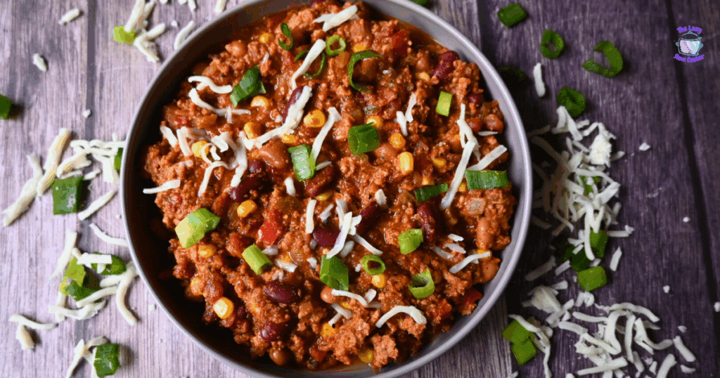 A bowl of chili with ground turkey, beans, corn, tomatoes, topped with shredded cheese and sliced green onions, sits on a wooden surface with extra cheese and onions scattered around.
