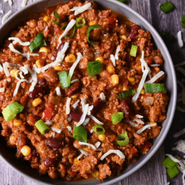 A bowl of chili with ground turkey, beans, corn, tomatoes, topped with shredded cheese and sliced green onions, sits on a wooden surface with extra cheese and onions scattered around.