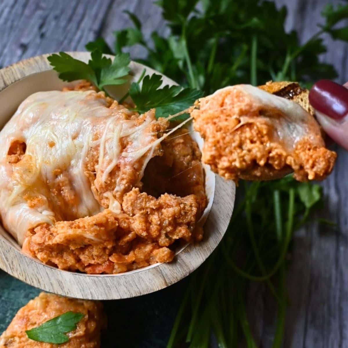 cheesy chicken parm dip on a cracker being held over a bowl full of the same