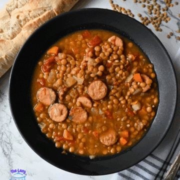 A black bowl filled with lentil soup containing sliced sausage, carrots, onions, and tomatoes sits next to a baguette and scattered dry lentils on a marble surface. A striped napkin and spoon are nearby.