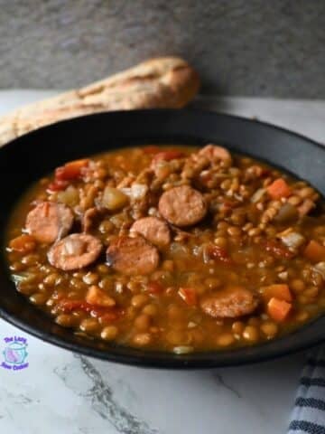 A black bowl filled with lentil soup and sliced sausage sits on a marble surface, with a piece of bread in the background and a striped cloth to the side.