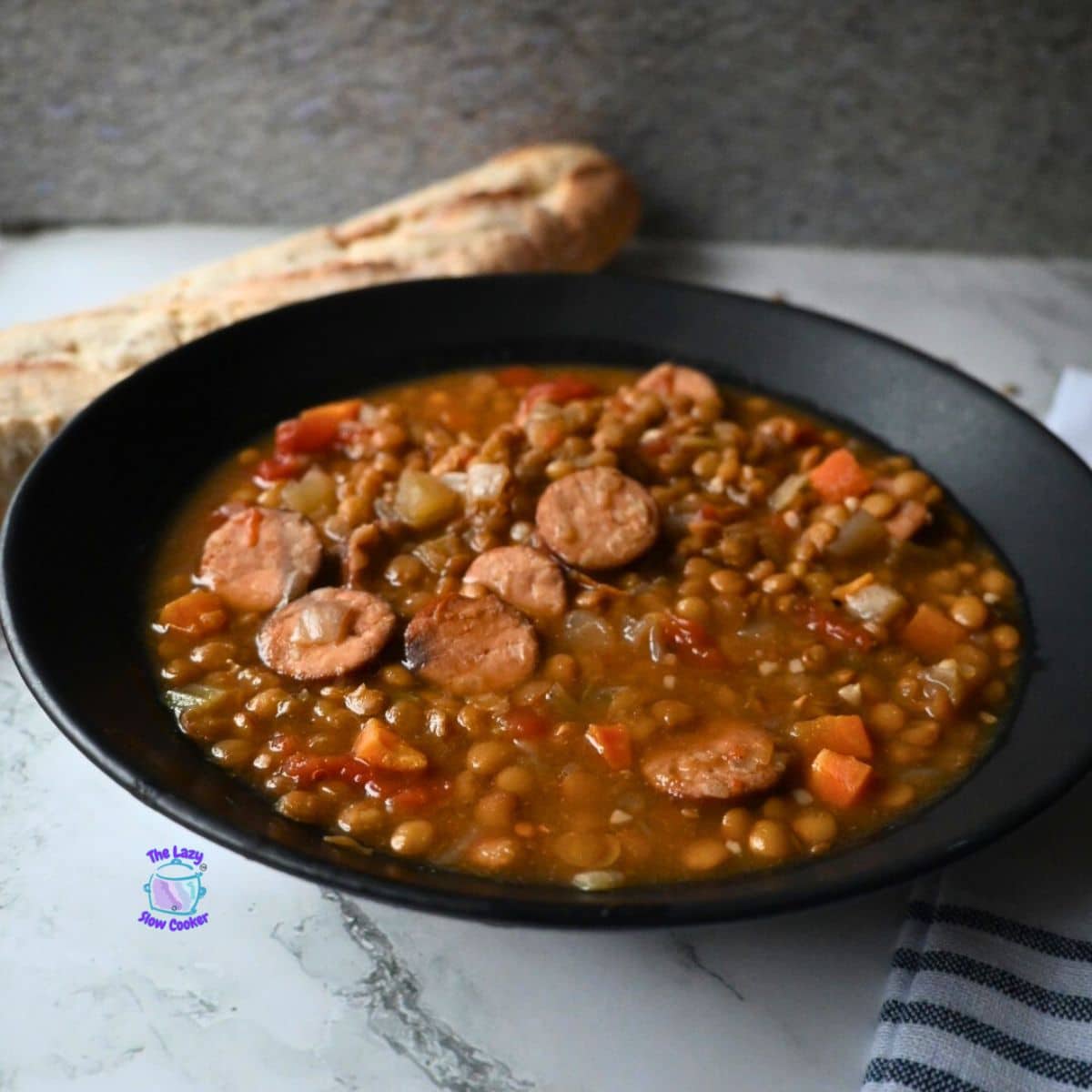 A black bowl filled with lentil soup and sliced sausage sits on a marble surface, with a piece of bread in the background and a striped cloth to the side.