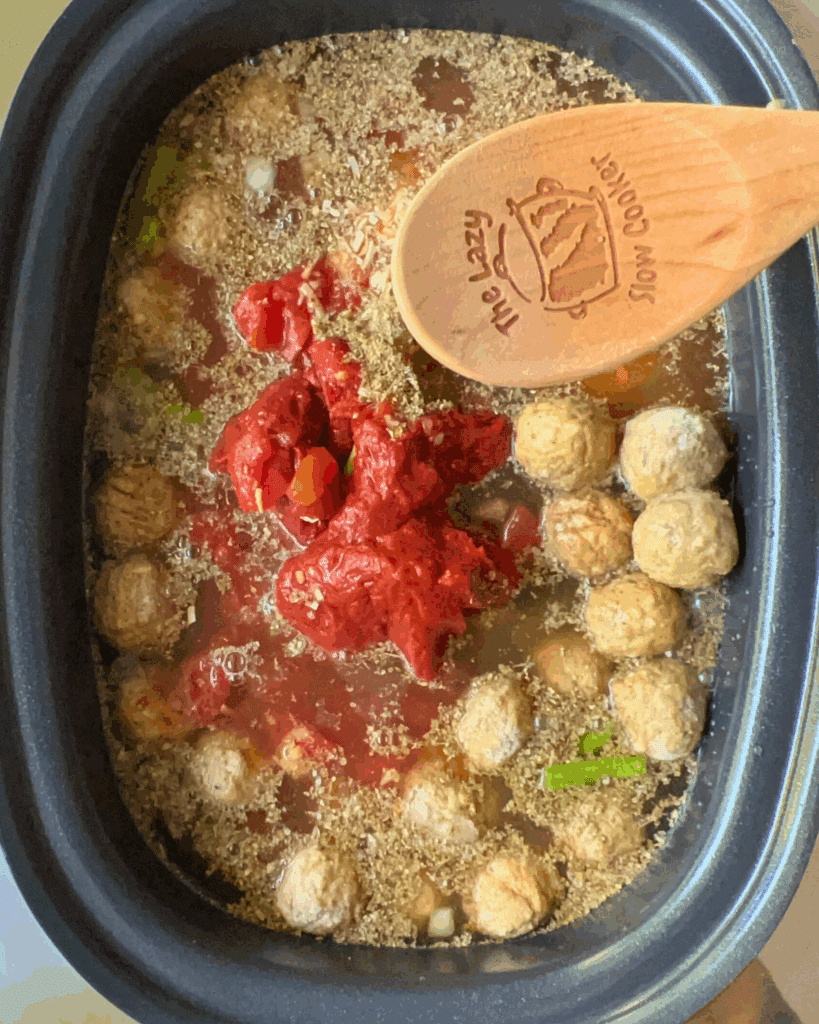 vegetables, meatballs, tomato paste, broth, and seasonings in slow cooker with a wooden spoon