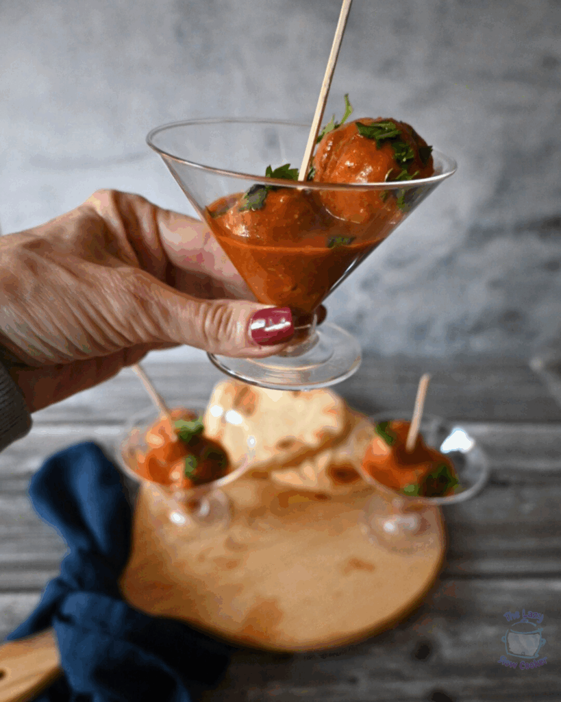 A hand holds a martini glass containing two skewered meatballs in red sauce, garnished with herbs. Two similar glasses and pieces of flatbread are on a wooden board in the background.