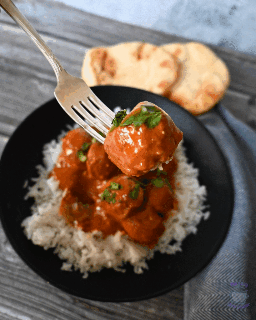 A fork holds a saucy meatball above a black bowl filled with white rice and more meatballs in sauce, garnished with herbs. Two pieces of naan bread are visible in the background.