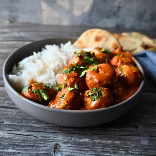 A bowl of white rice and butter chicken sauce with chicken meatballs, garnished with chopped herbs, served with naan bread on a wooden surface next to a blue-striped napkin.