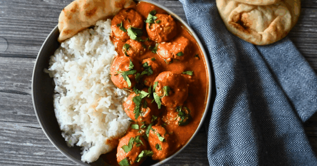 A gray plate with white rice slow cooker butter chicken meatballs in a creamy orange sauce, garnished with chopped cilantro, next to pieces of naan bread and a blue textured cloth