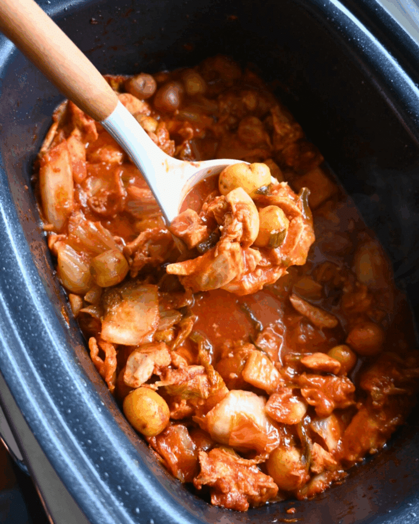 A slow cooker filled with a hearty stew featuring chunks of chicken, baby potatoes, vegetables, and a rich red tomato-based sauce. A white ladle is scooping out a serving.