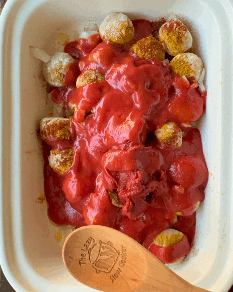 Chicken meatballs in a casserole style slow cooker are topped with red tomato sauce and spices, ready to be cooked. A wooden spoon engraved with The Lazy Slow Cooker rests nearby.