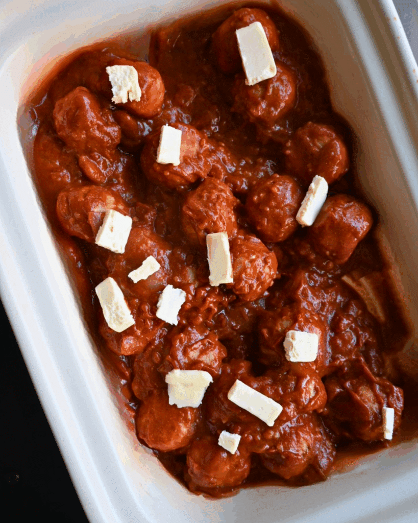 Chicken meatballs covered in sauce topped with several small cubes of butter in a white rectangular slow cooker.