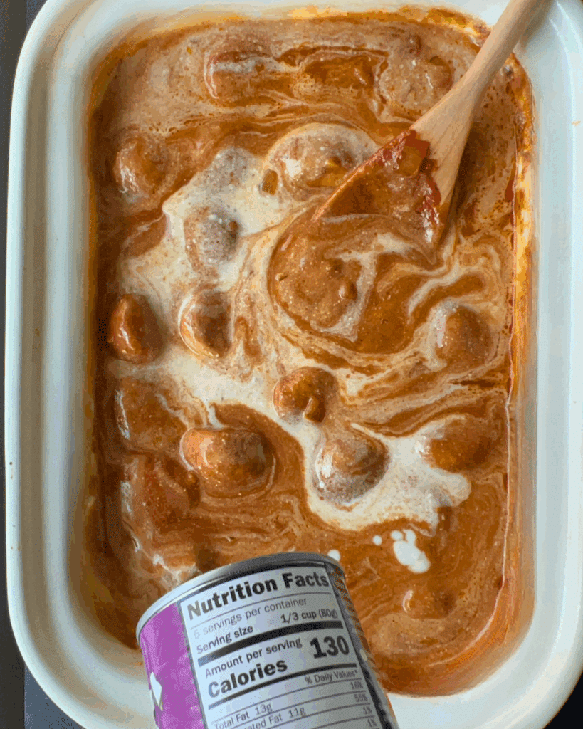 A slow-cooked butter chicken meatballs dish in creamy sauce being stirred with a wooden spoon, with a can of coconut milk visible at the bottom edge