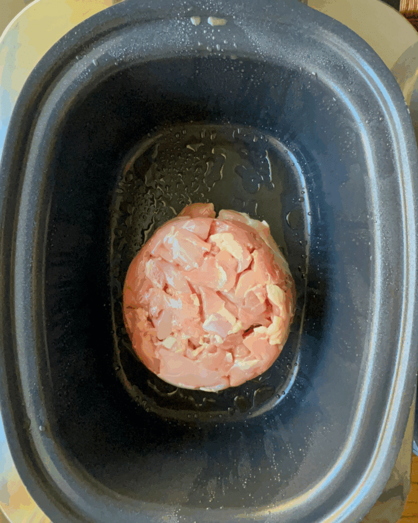Raw, diced chicken formed into a round mound sits in the center of a black slow cooker insert, with a light coating of oil around it.