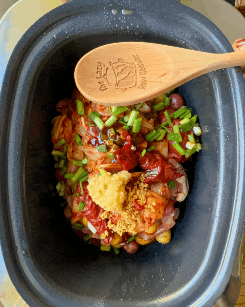A slow cooker filled with raw ingredients, including chopped green onions, kimchi, minced garlic, sauce, and meat, topped with a wooden spoon that reads