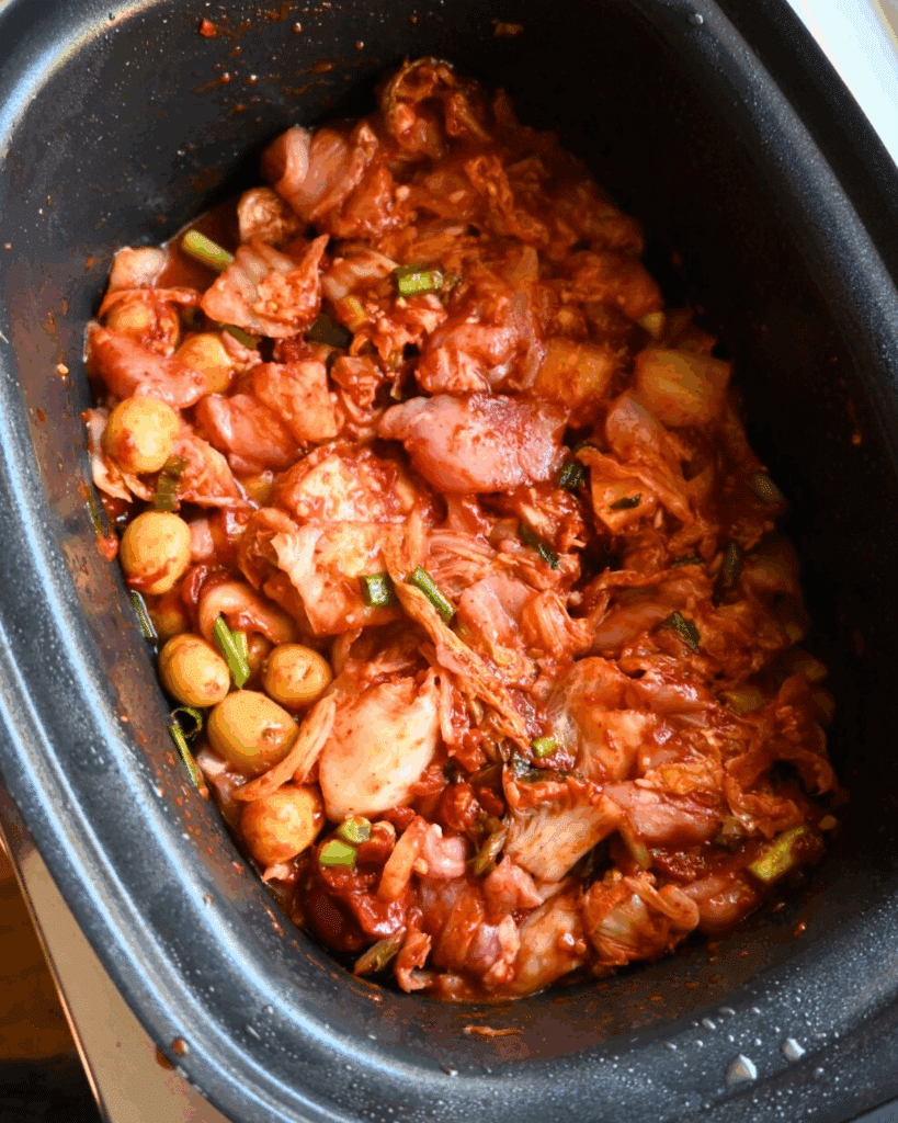 Marinated kimchi mixed with vegetables, including cabbage, green onions, and small mushrooms, sits in a black rectangular slow cooker ready for fermentation or cooking.