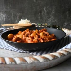 A black bowl filled with a red, saucy slow cooker Kimchi chicken and potato dish sits on a striped cloth, with rice, chopsticks, and seaweed in the background. The setting appears rustic with a textured dark backdrop.