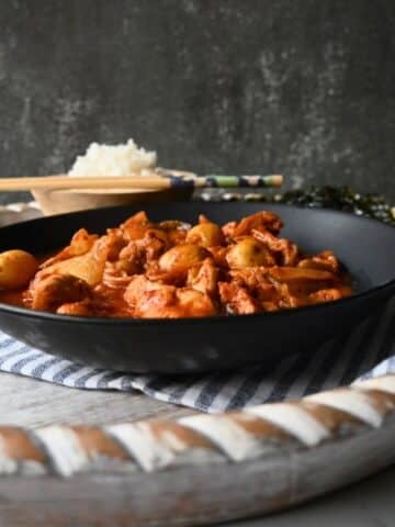 A black bowl filled with a red, saucy slow cooker Kimchi chicken and potato dish sits on a striped cloth, with rice, chopsticks, and seaweed in the background. The setting appears rustic with a textured dark backdrop.