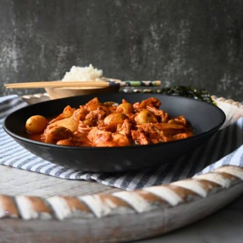 A black bowl filled with a red, saucy slow cooker Kimchi chicken and potato dish sits on a striped cloth, with rice, chopsticks, and seaweed in the background. The setting appears rustic with a textured dark backdrop.
