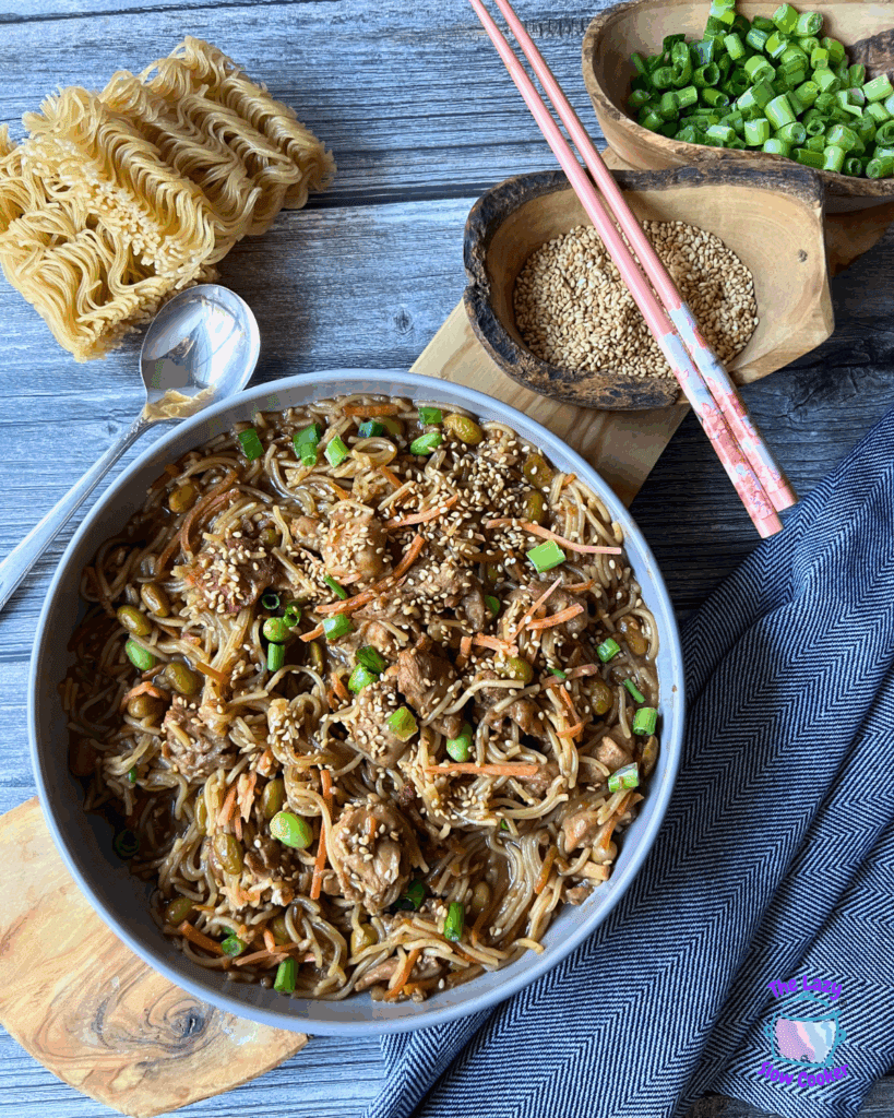A bowl of slow cooked sesame chicken and noodles with chicken, carrot strips, chopped green onions, and sesame seeds, with chopsticks resting on top. A small bowl of sesame seeds is beside the main dish on a wooden board.