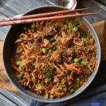 A bowl of slow cooked sesame chicken and noodles with chicken, carrot strips, chopped green onions, and sesame seeds, with chopsticks resting on top. A small bowl of sesame seeds is beside the main dish on a wooden board.
