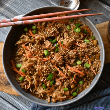 A bowl of slow cooked sesame chicken and noodles with chicken, carrot strips, chopped green onions, and sesame seeds, with chopsticks resting on top. A small bowl of sesame seeds is beside the main dish on a wooden board.
