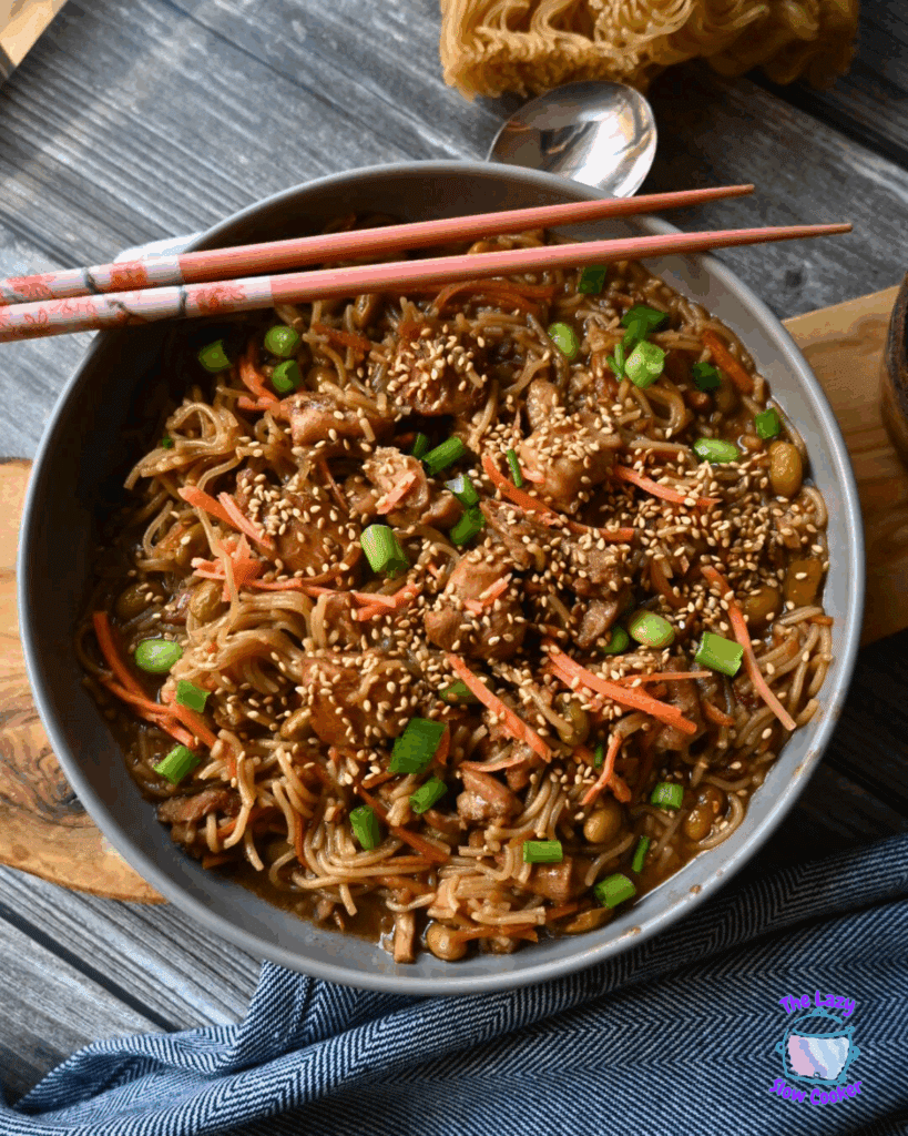 A bowl of slow cooked sesame chicken and noodles with chicken, carrot strips, chopped green onions, and sesame seeds, with chopsticks resting on top. A small bowl of sesame seeds is beside the main dish on a wooden board.
