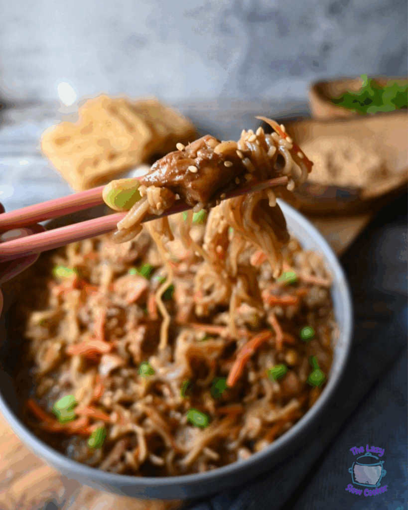 Close-up of chopsticks holding a piece of food coated in sesame seeds above a bowl of stir-fried noodles with vegetables. Scattered green onions and sesame seeds are on a rustic wooden surface.