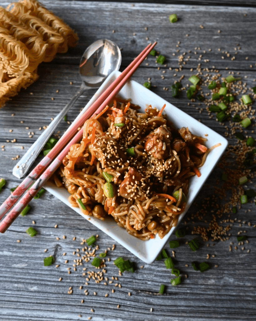 A square white bowl filled with sesame chicken and noodles, topped with sesame seeds and green onions, sits on a wooden table with chopsticks and a roll in the background. Slow Cooker Sesame Chicken is written on the image.