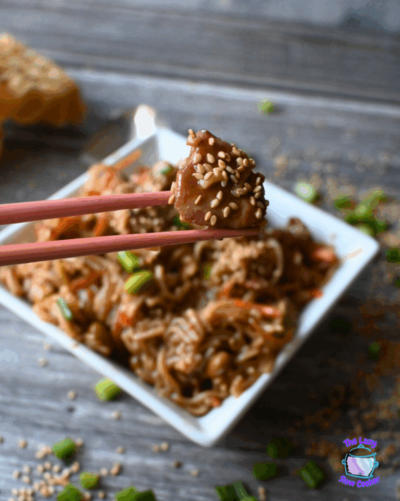 Close-up of chopsticks holding a piece of chicken coated in sesame seeds above a bowl of slow cooked noodles with vegetables. Scattered green onions and sesame seeds are on a rustic wooden surface.