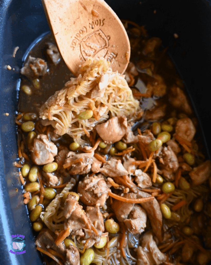 A wooden spoon lifts ramen noodles in a dark sesame sauce with carrots, chicken and edamame from a black slow cooker. The mixture appears hearty and well-cooked.
