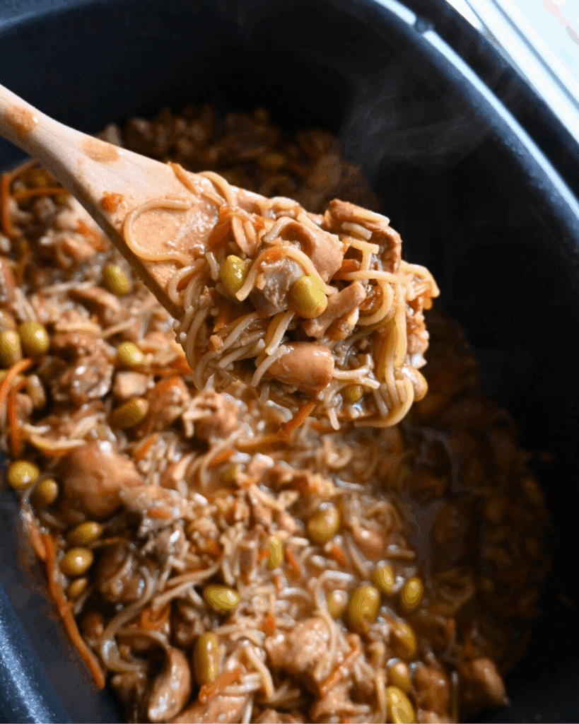 A wooden spoon lifts ramen noodles in a dark sesame sauce with carrots, chicken and edamame from a black slow cooker. The mixture appears hearty and well-cooked.