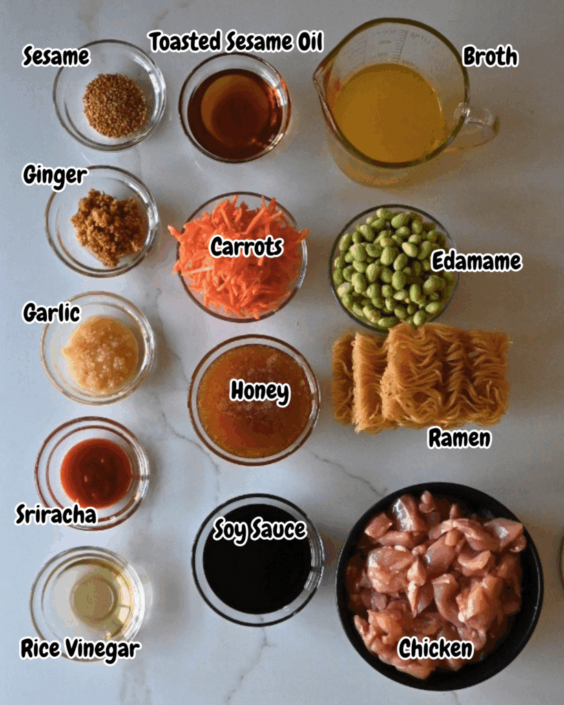 Various labeled ingredients for a slow cooked chicken and noodle dish are arranged on a counter, including chicken, ramen noodles, edamame, carrots, broth, toasted sesame oil, sesame seeds, ginger, garlic, honey, sriracha, soy sauce, and rice vinegar.