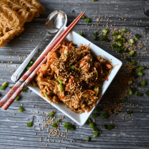 A square white bowl filled with sesame chicken and noodles, topped with sesame seeds and green onions, sits on a wooden table with chopsticks and a roll in the background. Slow Cooker Sesame Chicken is written on the image.