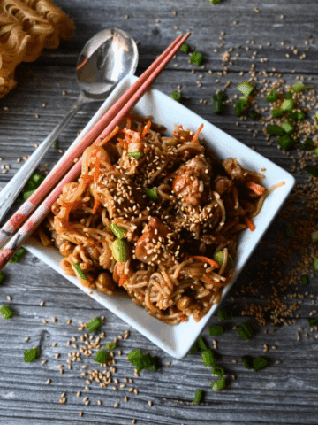 A square white bowl filled with sesame chicken and noodles, topped with sesame seeds and green onions, sits on a wooden table with chopsticks and a roll in the background. Slow Cooker Sesame Chicken is written on the image.