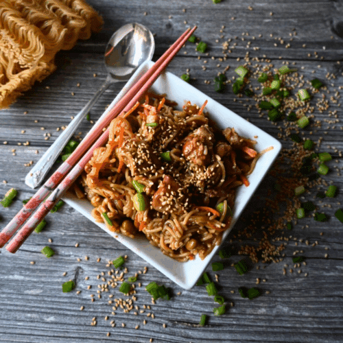 A square white bowl filled with sesame chicken and noodles, topped with sesame seeds and green onions, sits on a wooden table with chopsticks and a roll in the background. Slow Cooker Sesame Chicken is written on the image.