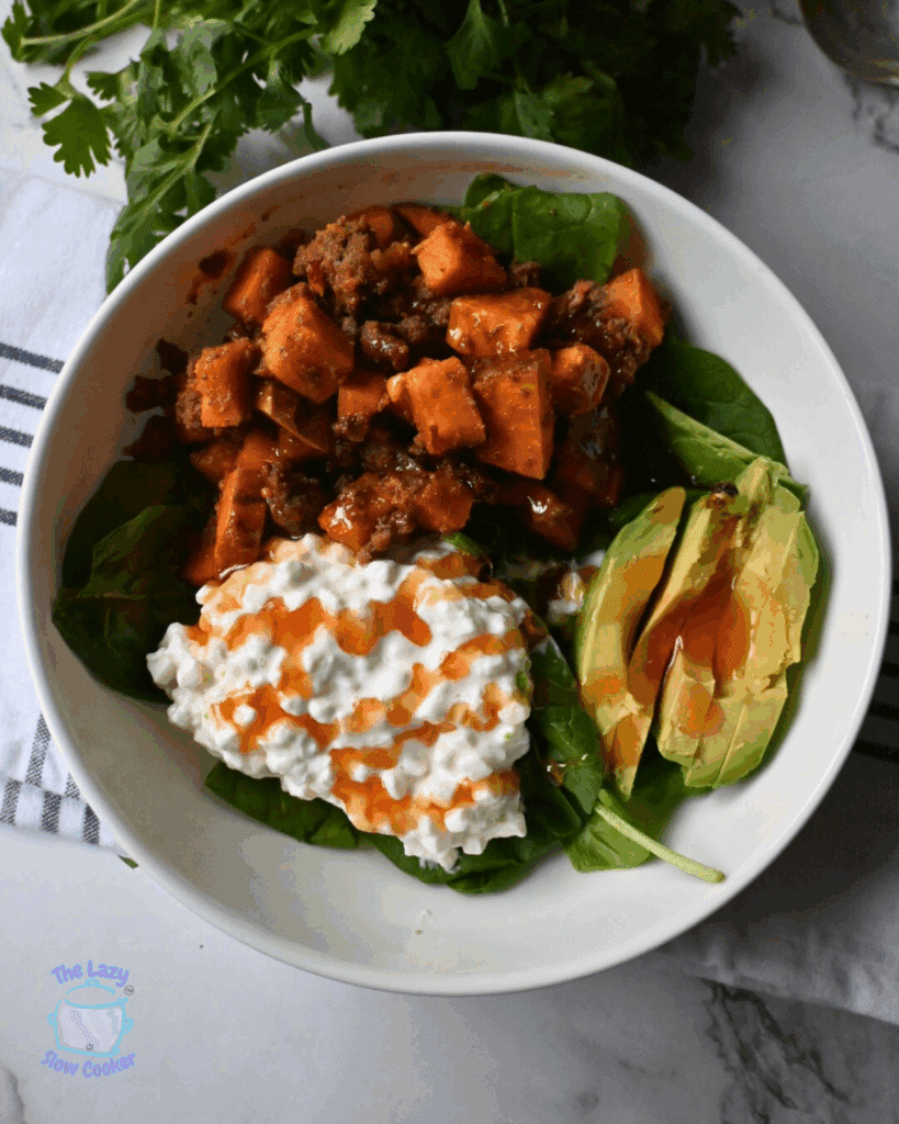A viral beef and cottage cheese protein bowl containing spinach, cottage cheese drizzled with hot honey sauce, sliced avocado, and a sweet potato and ground meat mixture, all placed on a gray bowl