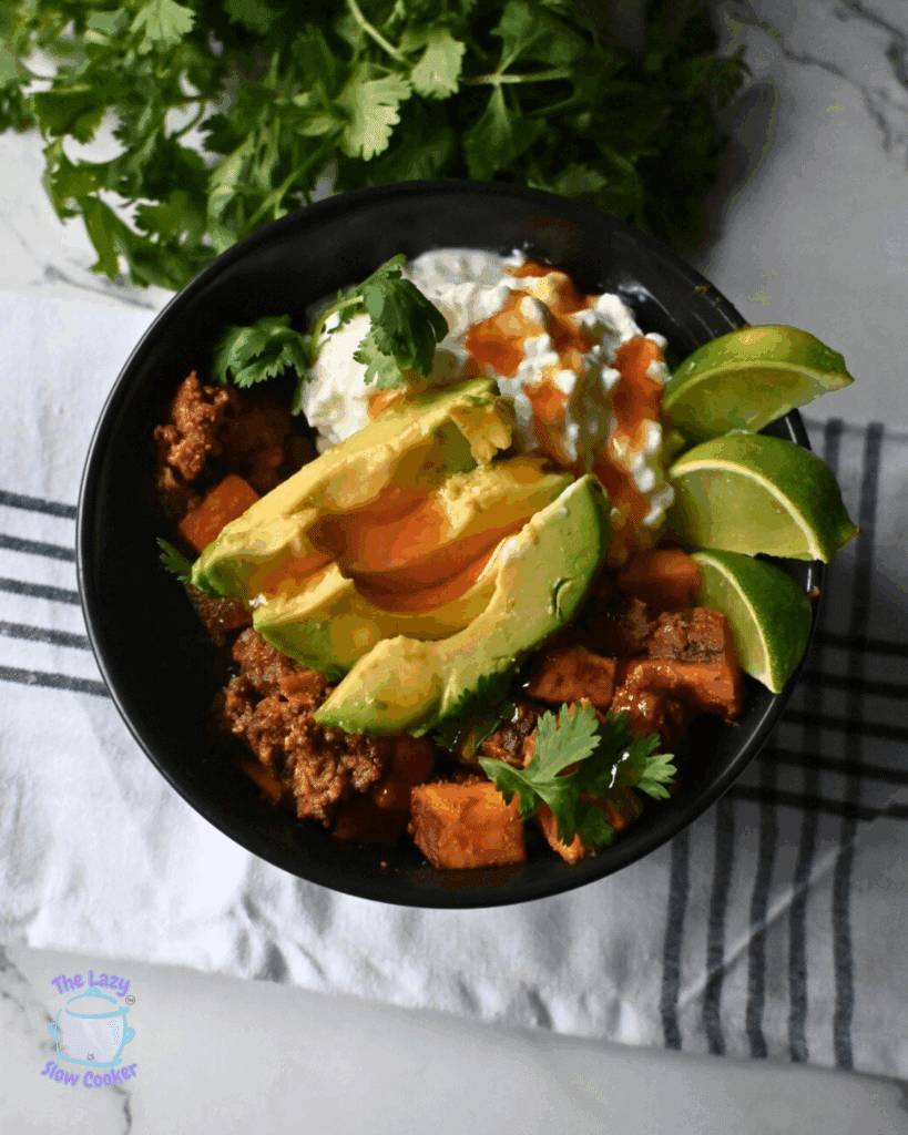 A black bowl filled with taco ground beef, avocado slices, lime wedges, cottage cheese, hot sauce, and cilantro, placed on a striped kitchen towel with fresh cilantro in the background.