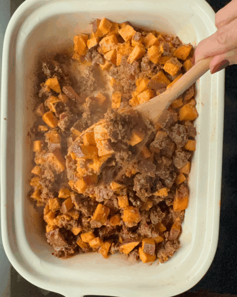 A hand stirs a mixture of slow cooker cooked ground beef and diced sweet potatoes in a white baking dish with a wooden spoon. Steam is rising from the food, indicating it is hot and freshly made.