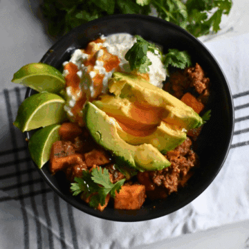 A black bowl filled with beef, sweet potatoes, avocado slices, cottage cheese, cilantro, and lime wedges sits on a striped cloth. The text Slow Cooker Viral Protein Beef Bowls appears at the bottom.