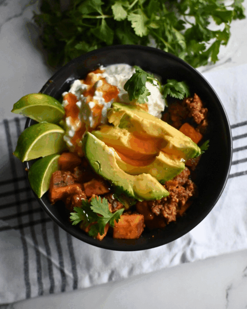 A black bowl filled with beef, sweet potatoes, avocado slices, cottage cheese, cilantro, and lime wedges sits on a striped cloth. The text Slow Cooker Viral Protein Beef Bowls appears at the bottom.