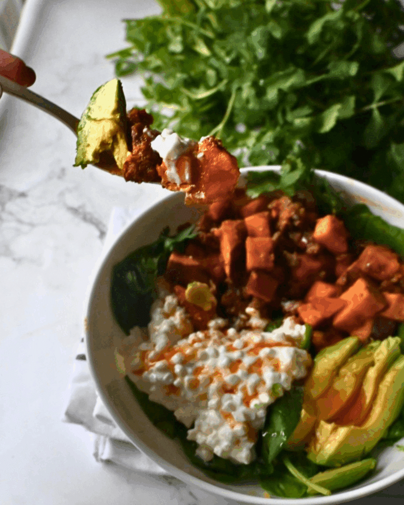 A close-up of a bowl with spinach, diced sweet potatoes, cottage cheese, avocado slices, and a drizzle of hot sauce. A hand holds a fork with a bite of avocado, sweet potato, and cottage cheese above the bowl. Fresh greens are in the background.