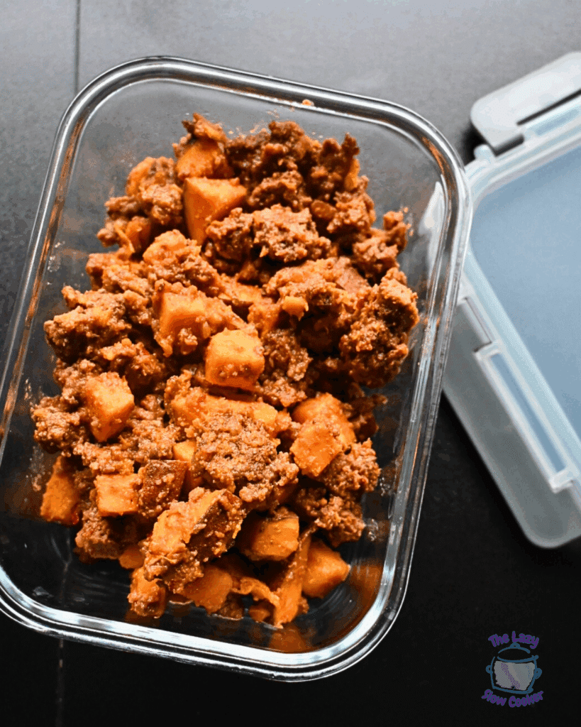 A glass container filled with slow cooked ground meat mixed with chunks of sweet potatoes, placed on a dark surface next to its partially open lid.