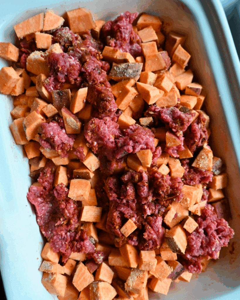 A casserole style slow cooker dish filled with cubed sweet potatoes and raw ground beef, seasoned and ready to be cooked.