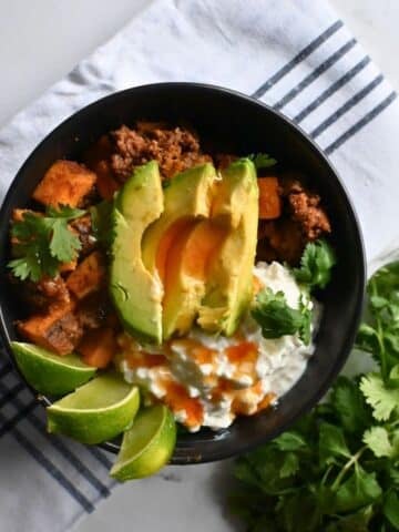 A black bowl filled with beef, sweet potatoes, avocado slices, cottage cheese, cilantro, and lime wedges sits on a striped cloth. The text Slow Cooker Viral Protein Beef Bowls appears at the bottom.