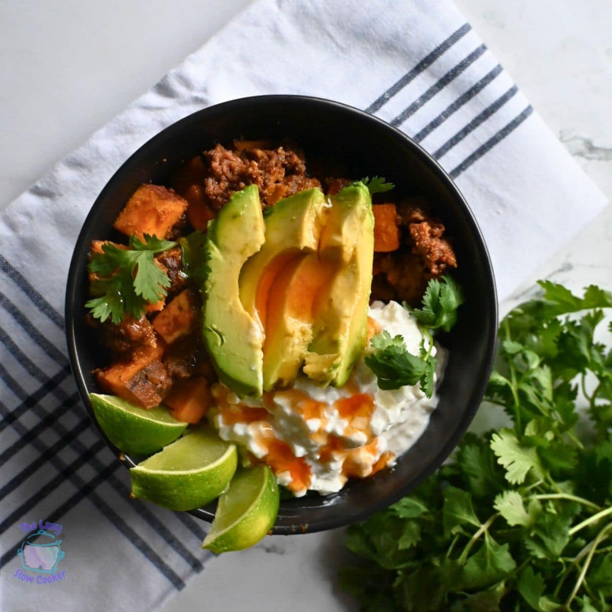 A black bowl filled with beef, sweet potatoes, avocado slices, cottage cheese, cilantro, and lime wedges sits on a striped cloth. The text Slow Cooker Viral Protein Beef Bowls appears at the bottom.